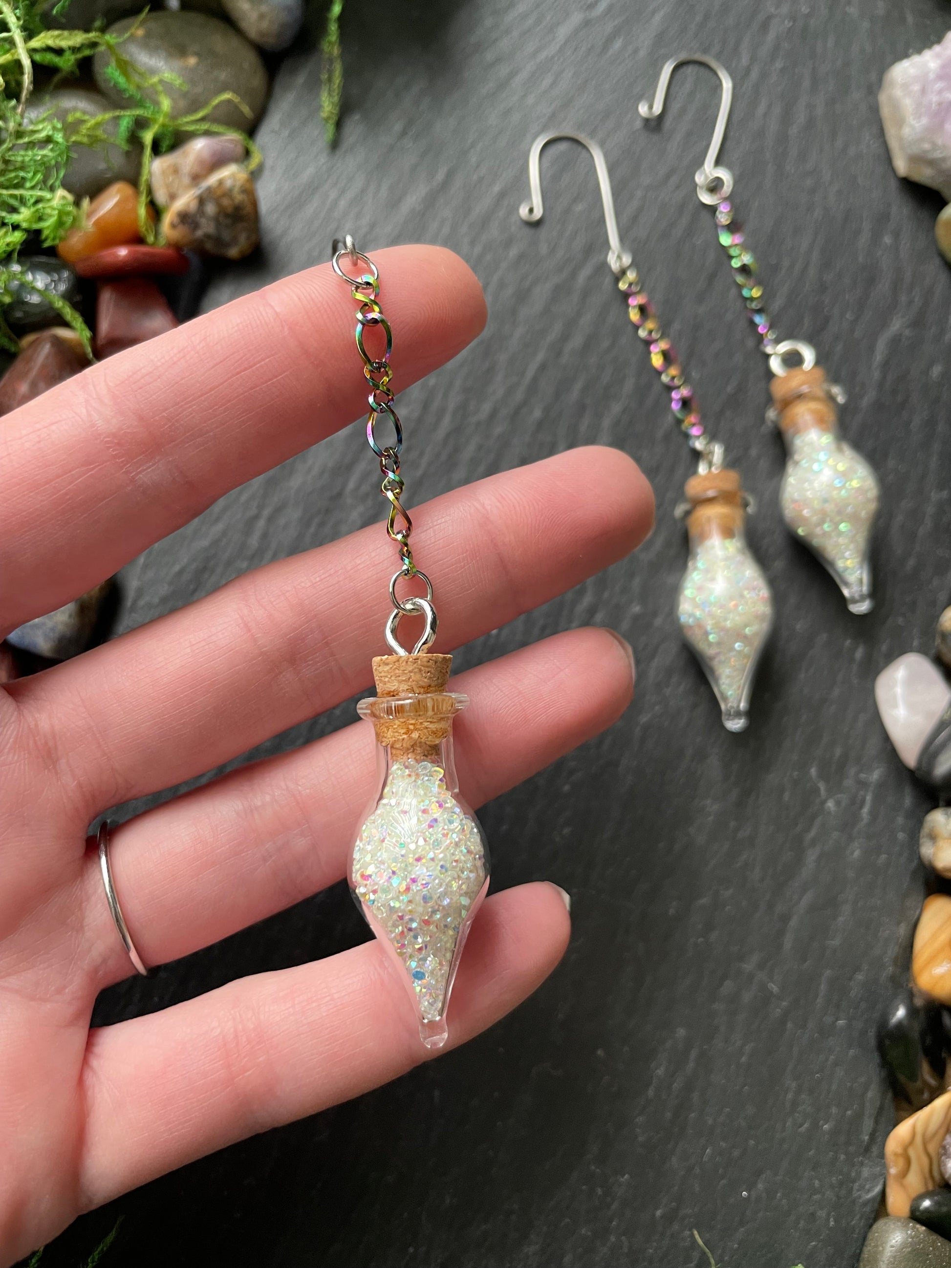 Hand holding decorative glass vials with sparkles on a dark surface with stones and plants.