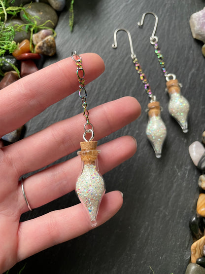 Hand holding decorative glass vials with sparkles on a dark surface with stones and plants.