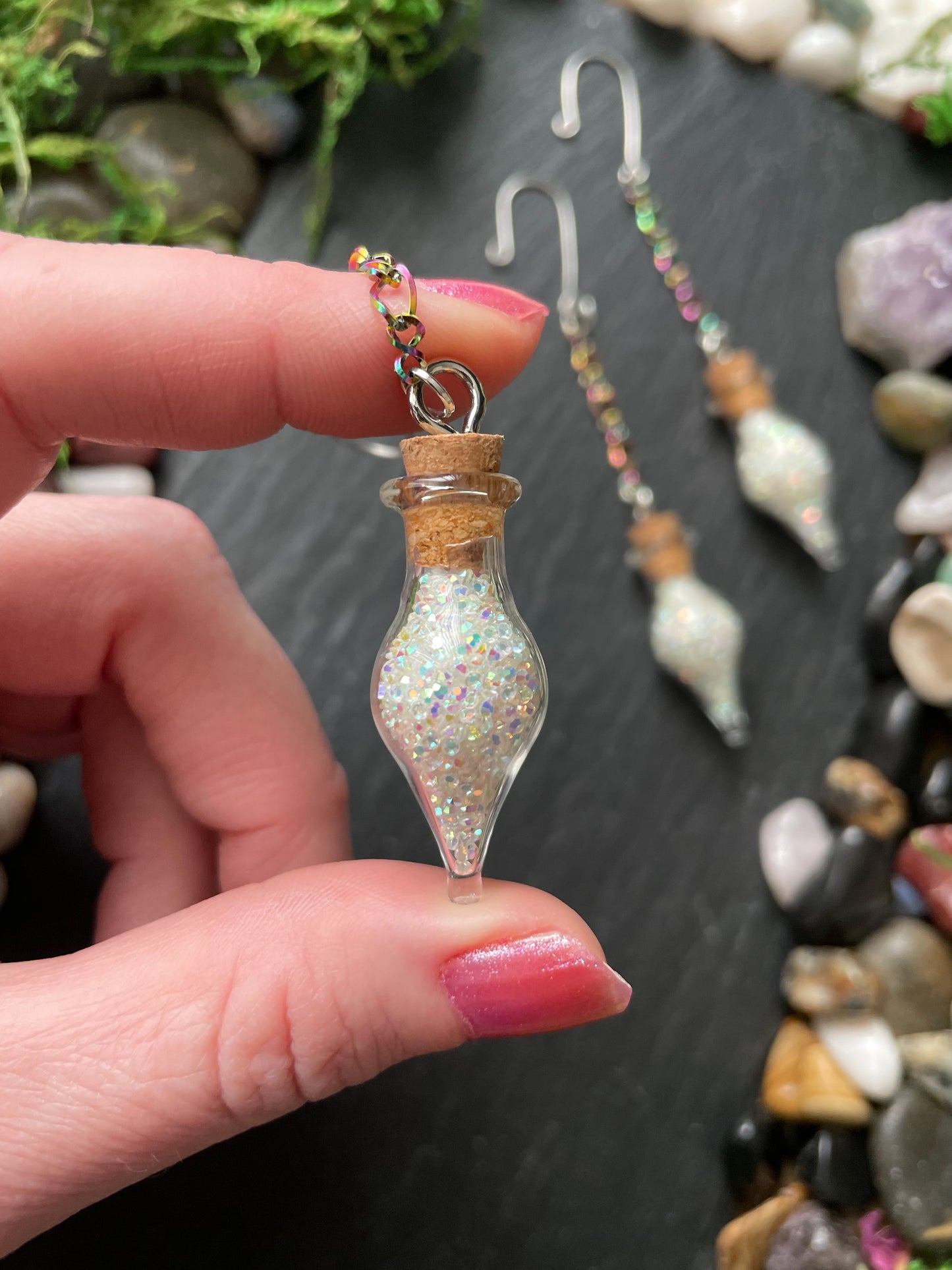 Hand holding a small glass vial charm with glittery contents against a backdrop of pebbles and stones.
