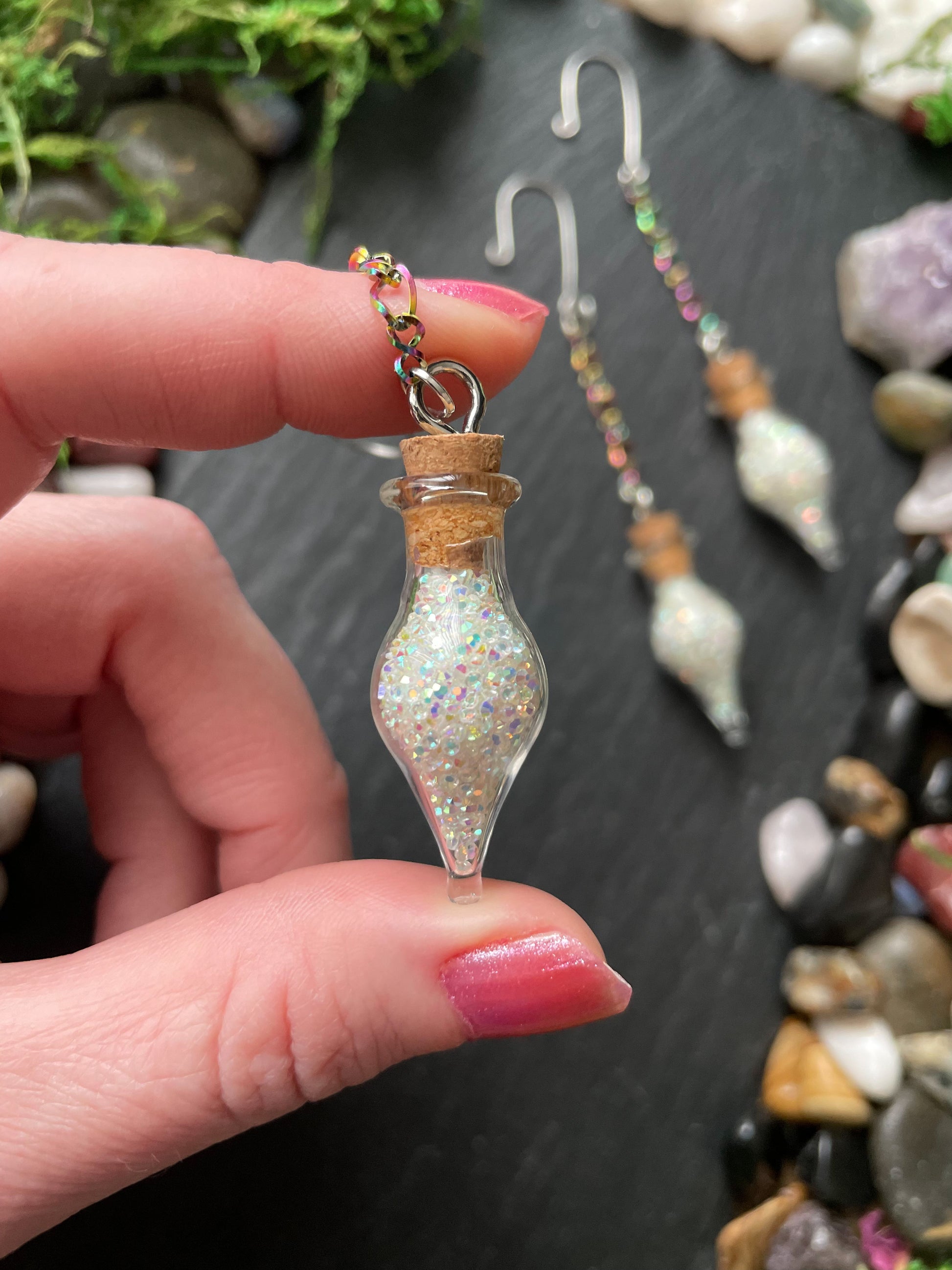 Hand holding a small glass vial charm with glittery contents against a backdrop of pebbles and stones.
