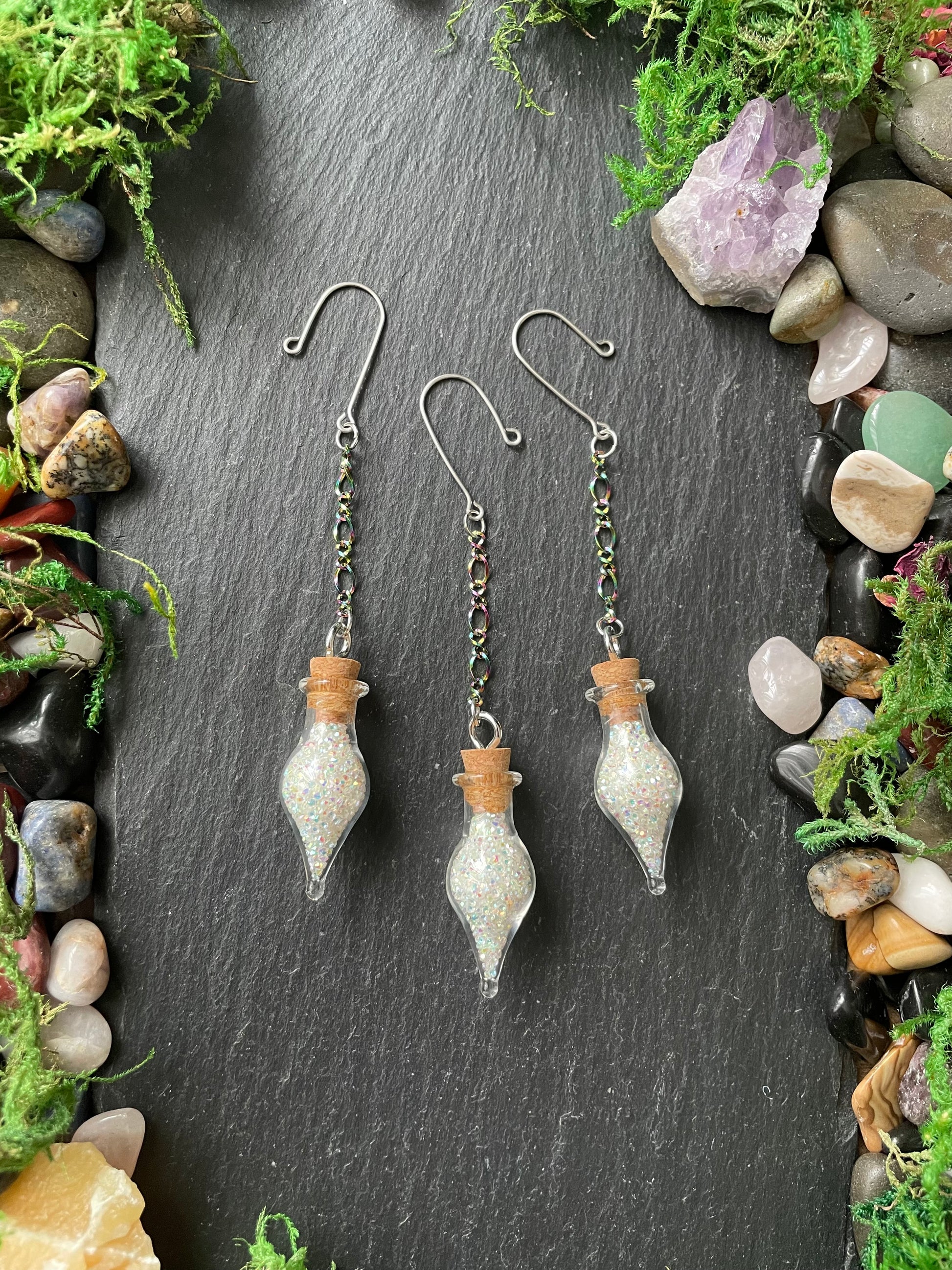Three glass bottle-shaped plant charms with cork stoppers on a dark stone surface surrounded by stones and greenery.