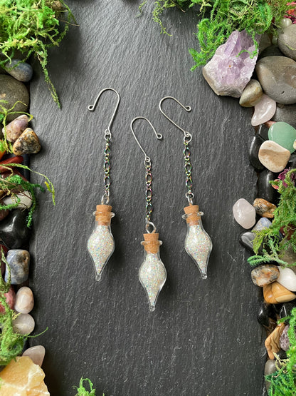 Three glass bottle-shaped plant charms with cork stoppers on a dark stone surface surrounded by stones and greenery.