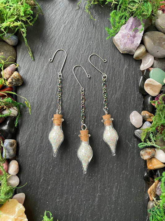 Three glass bottle-shaped plant charms with cork stoppers on a dark stone surface surrounded by stones and greenery.