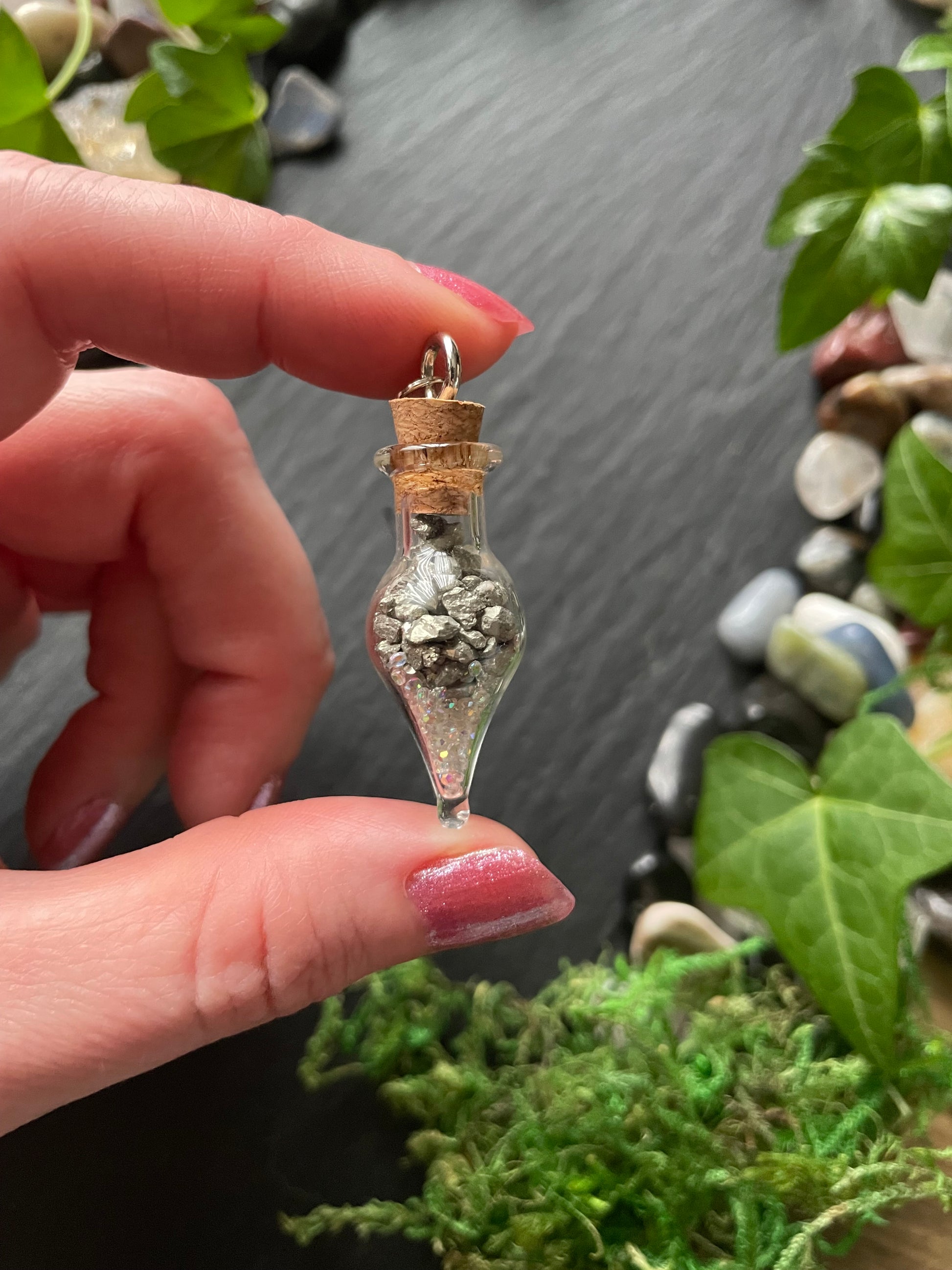 Hand holding a small decorative vial with pyrite crystals inside, against a natural background of stones and plants.