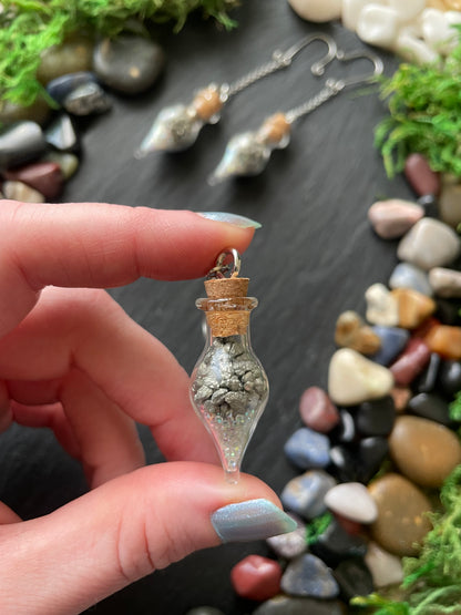 Hand holding a small glass vial with pyrite crystals against a stone and greenery background