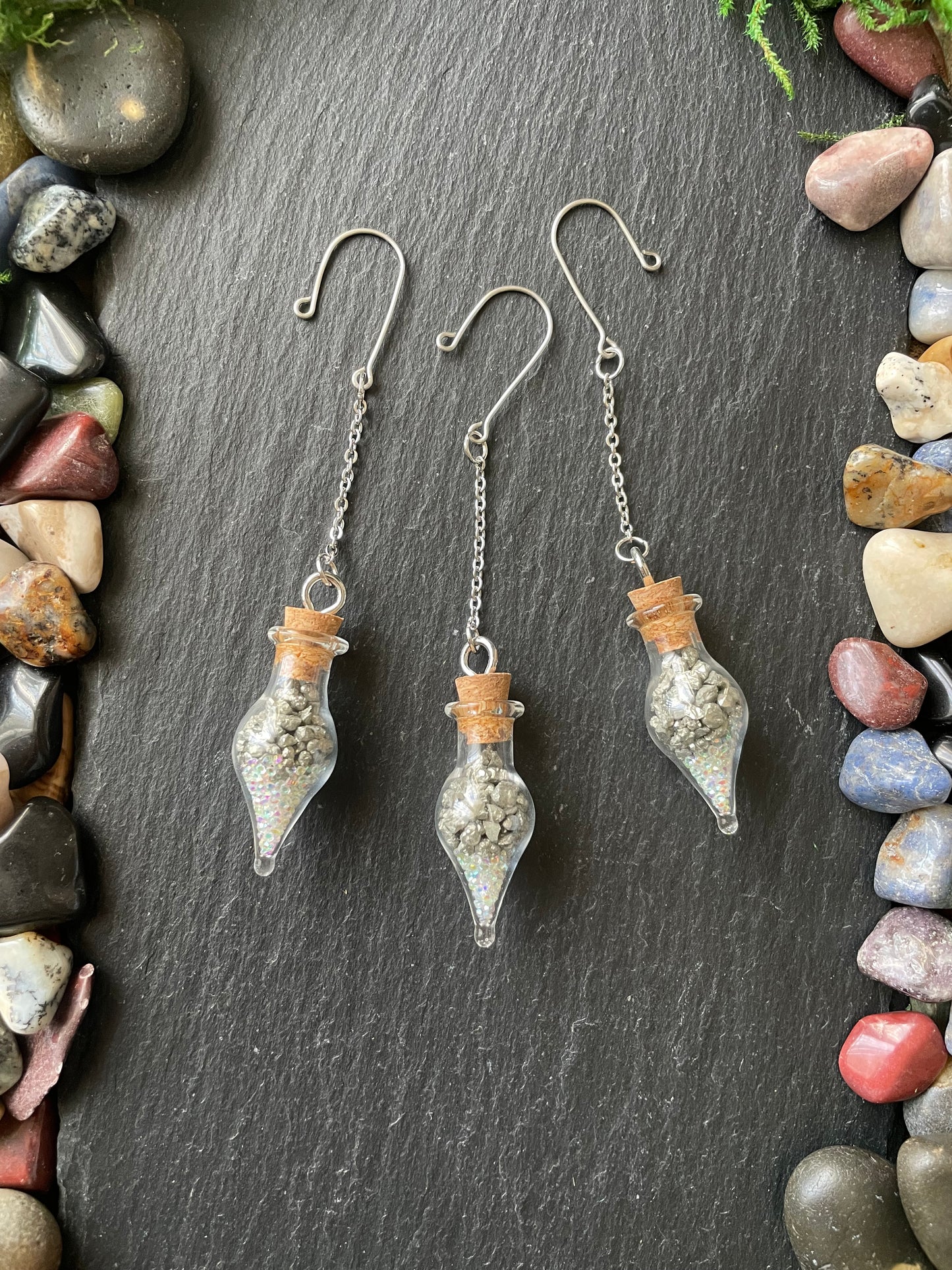 Set of glass teardrop-shaped vials with pyrite crystals and sparkles on a dark surface with pebbles around