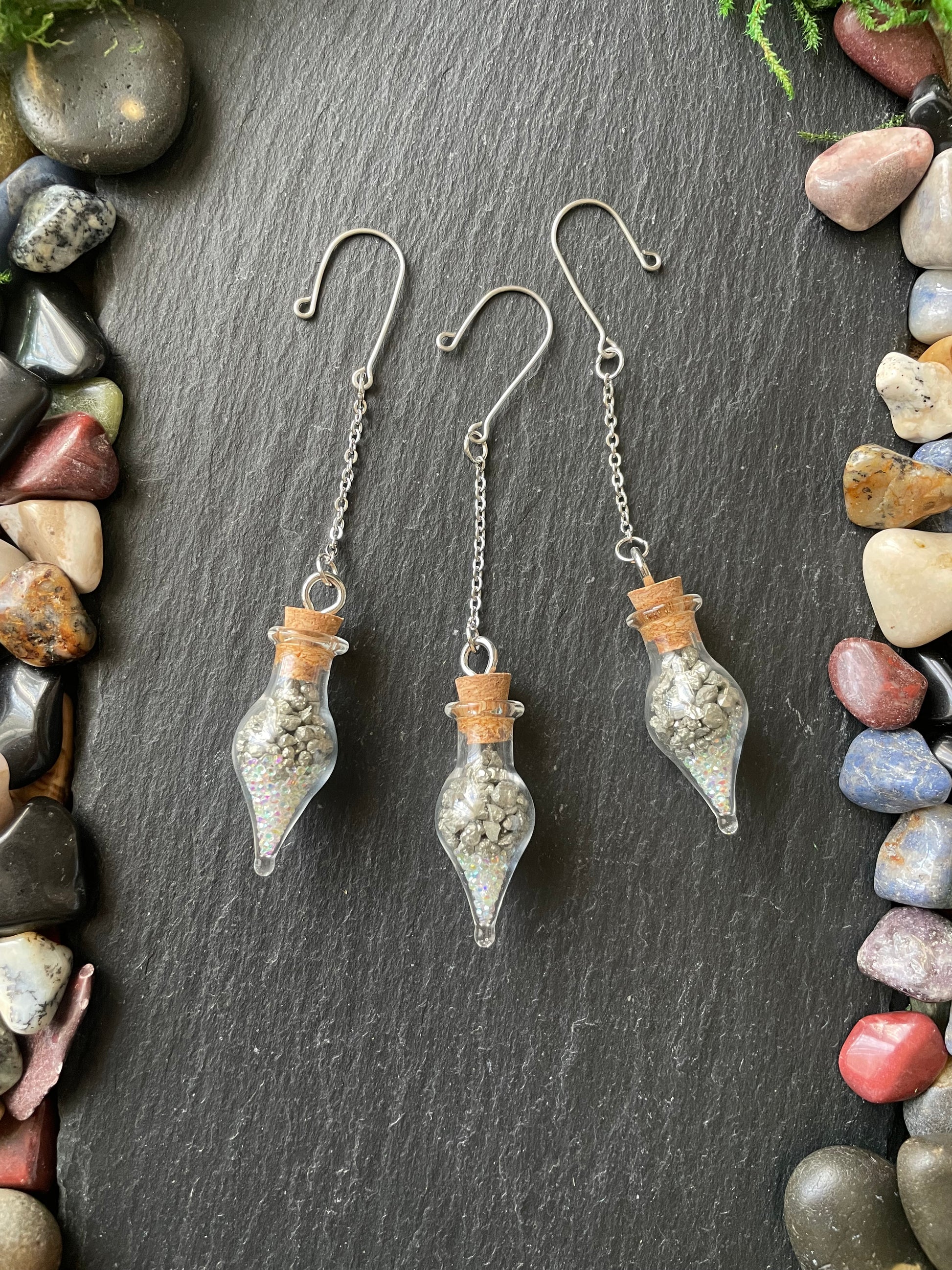 Set of glass teardrop-shaped vials with pyrite crystals and sparkles on a dark surface with pebbles around