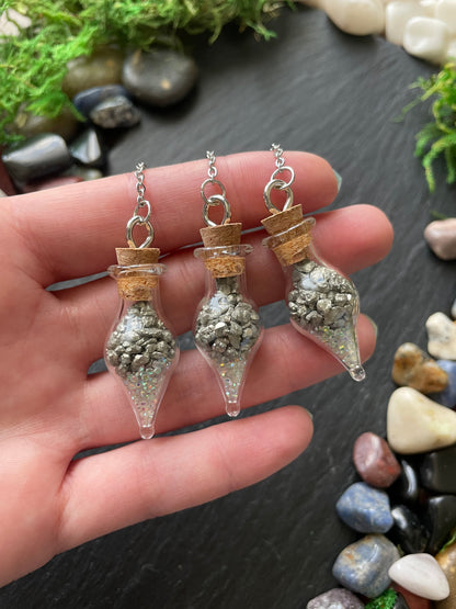 Hand holding three small decorative vials with crystals against a dark surface with stones and moss.