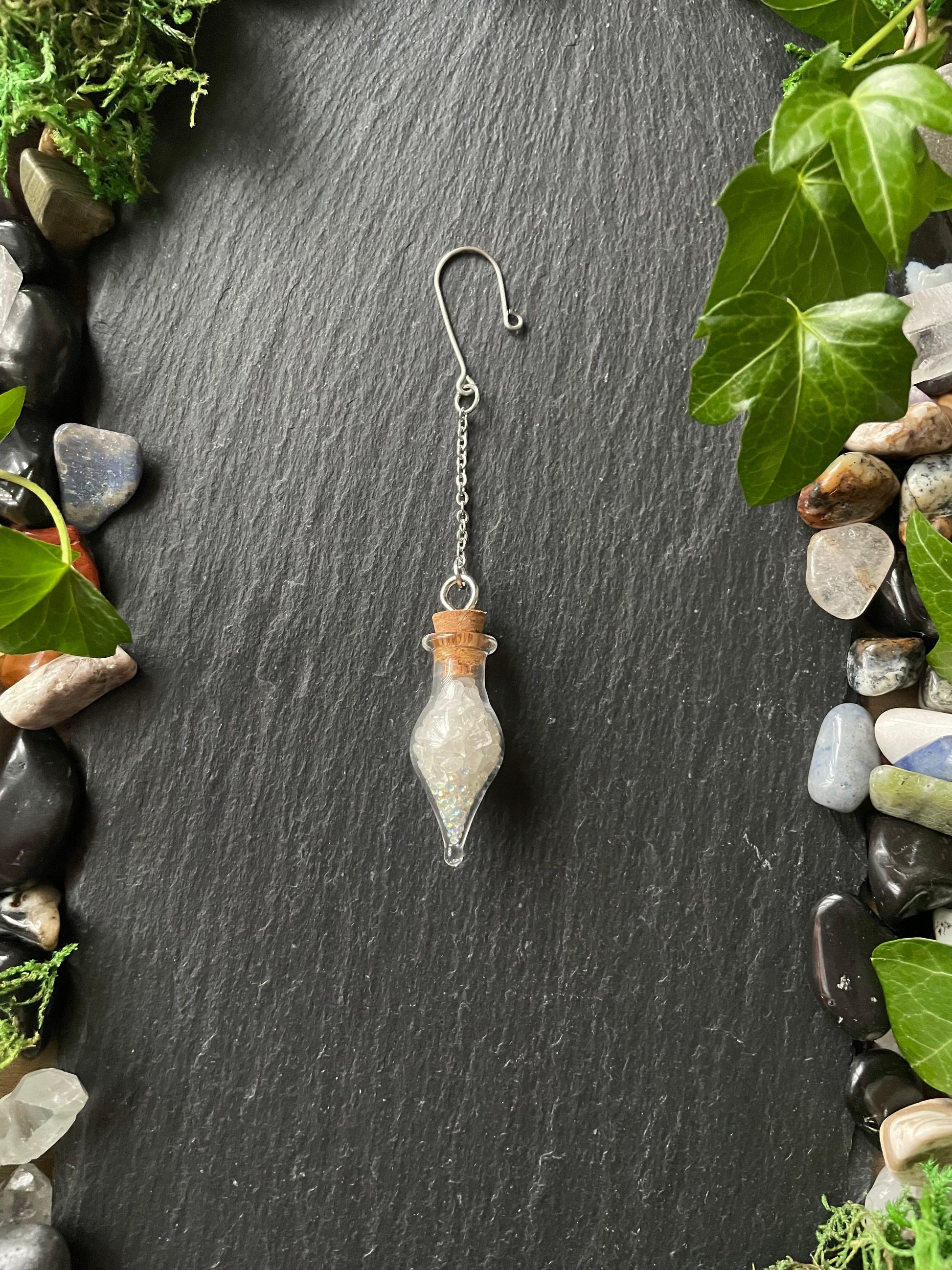 Decorative charm with a small glass container on a dark stone surface surrounded by greenery.