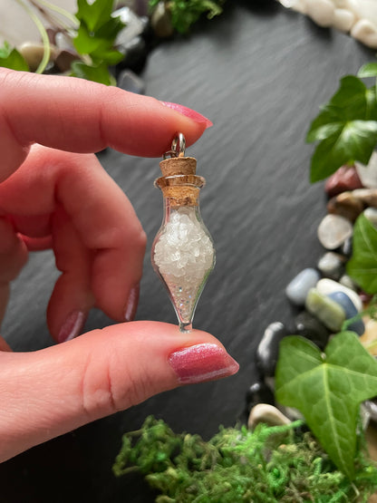 Hand holding a small decorative bottle with cork topper against a natural background of stones and plants.
