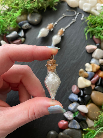 Hand holding a small glass vial with a cork against a background of stones and greenery