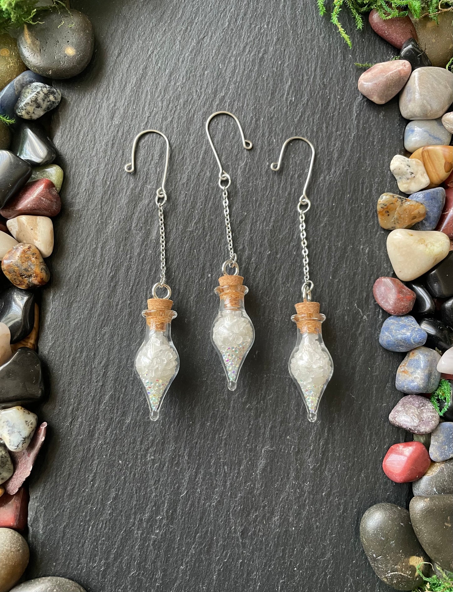 Three glass plant charms with cork stoppers on a dark slate surface surrounded by multicolored stones.