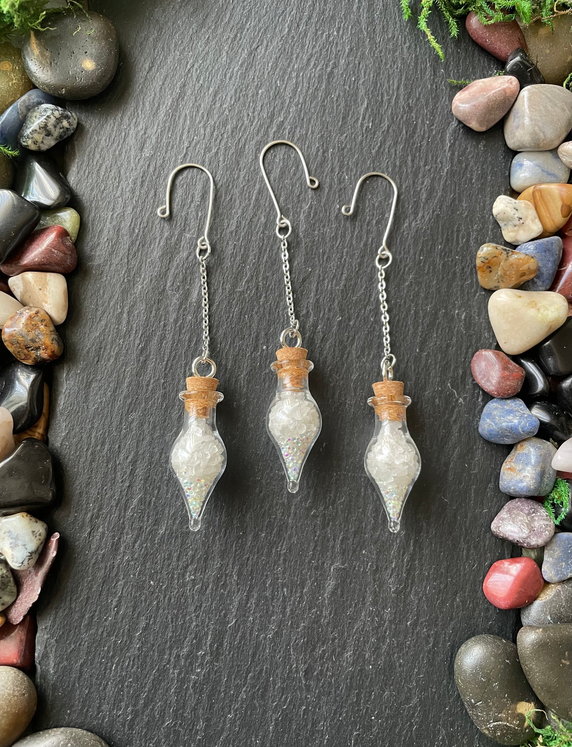 Three glass plant charms with cork stoppers on a dark slate surface surrounded by multicolored stones.