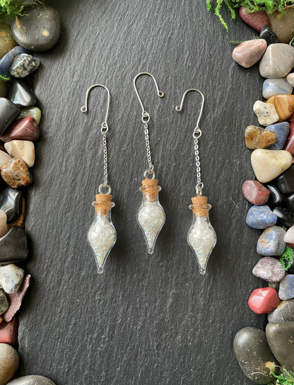Three glass plant charms with cork stoppers on a dark slate surface surrounded by multicolored stones.