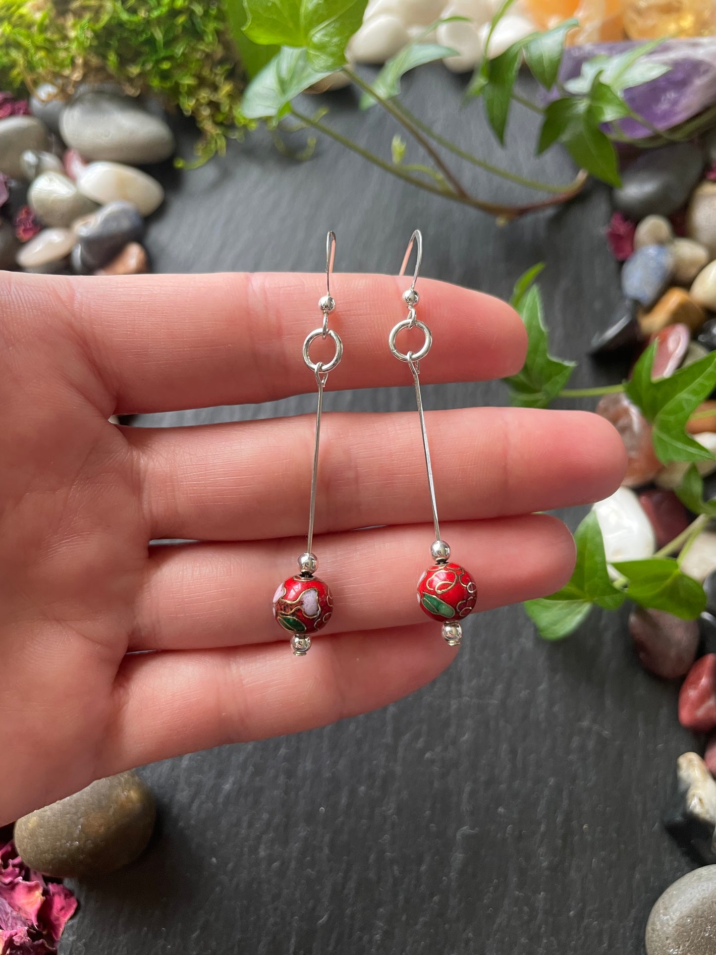 Silver earrings with red cloisonne beads on a hand against a natural background.
