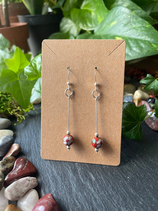 Silver earrings with red flower beads on a brown card with a natural background of stones and plants.
