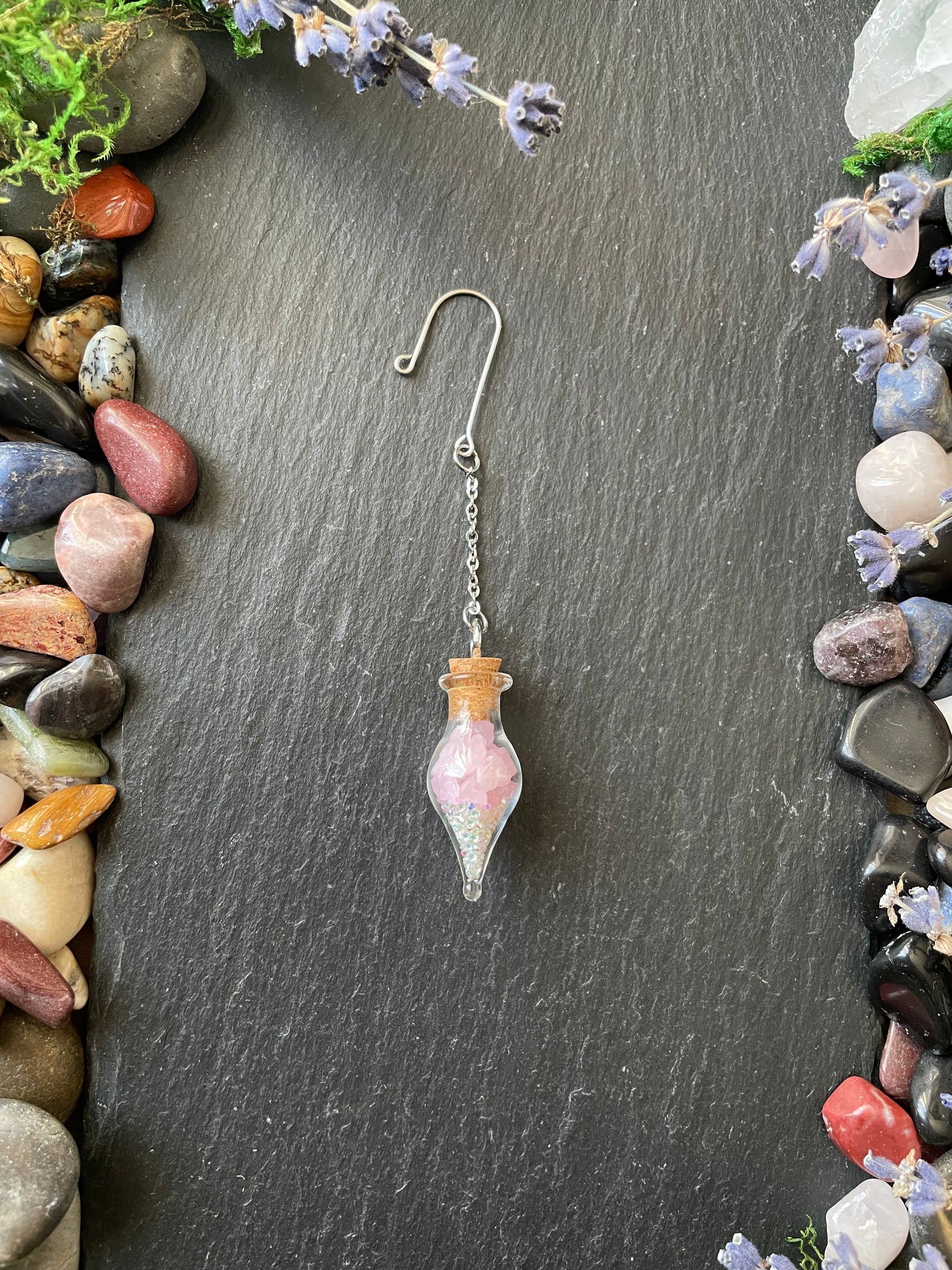 Glass vial with pink crystals and cork stopper on a dark surface surrounded by stones and plants