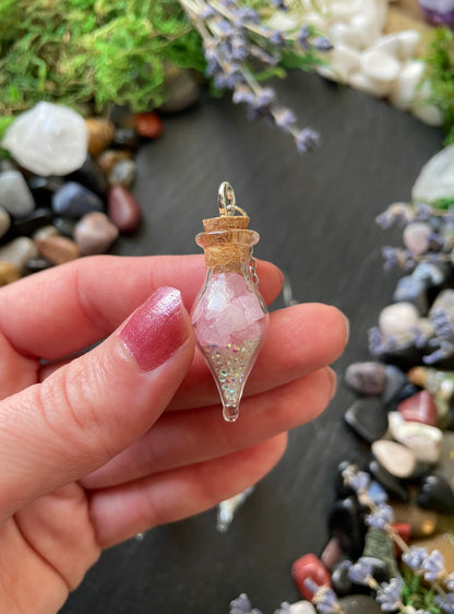 Hand holding a small glass vial with a pink crystals and sparkles inside against a natural background.