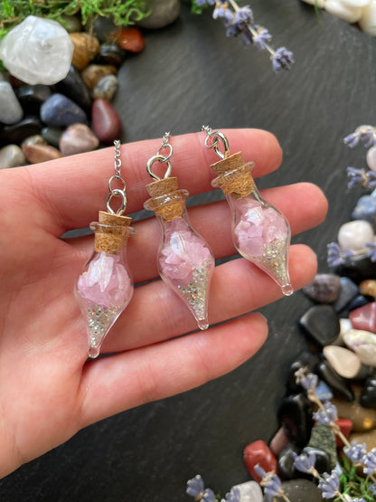 Three small glass bottle vials with pink crystals held in a hand against a natural stone background.