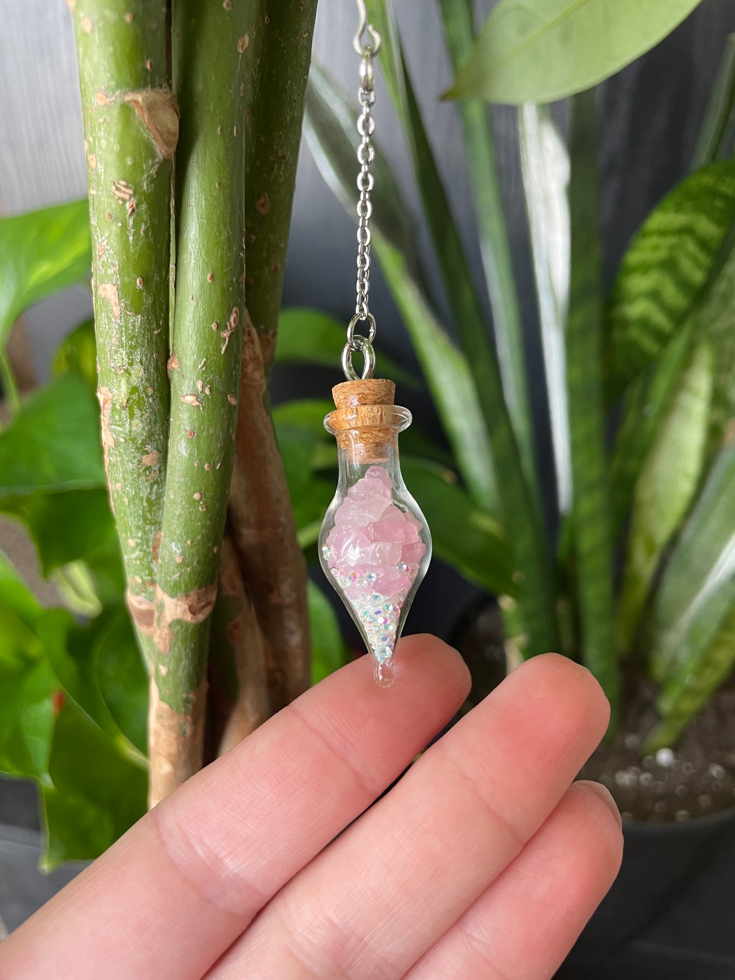 Hand holding a small glass bottle with pink crystals against a green leafy background