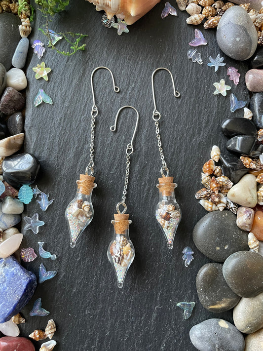 Glass bottle-shaped vials with seashells on a dark stone surface with pebbles and flowers.