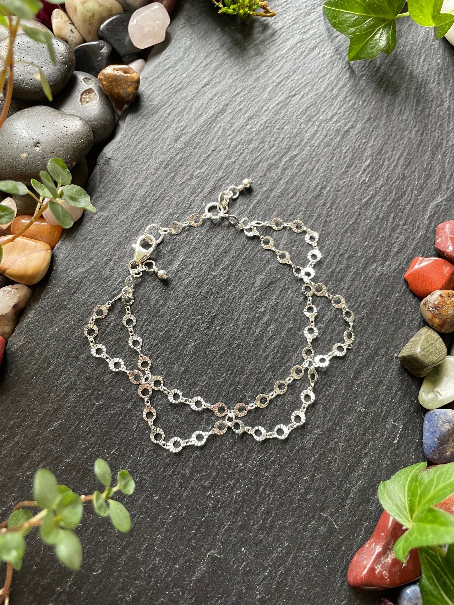 Silver chain bracelet on a dark stone surface with small stones and greenery around