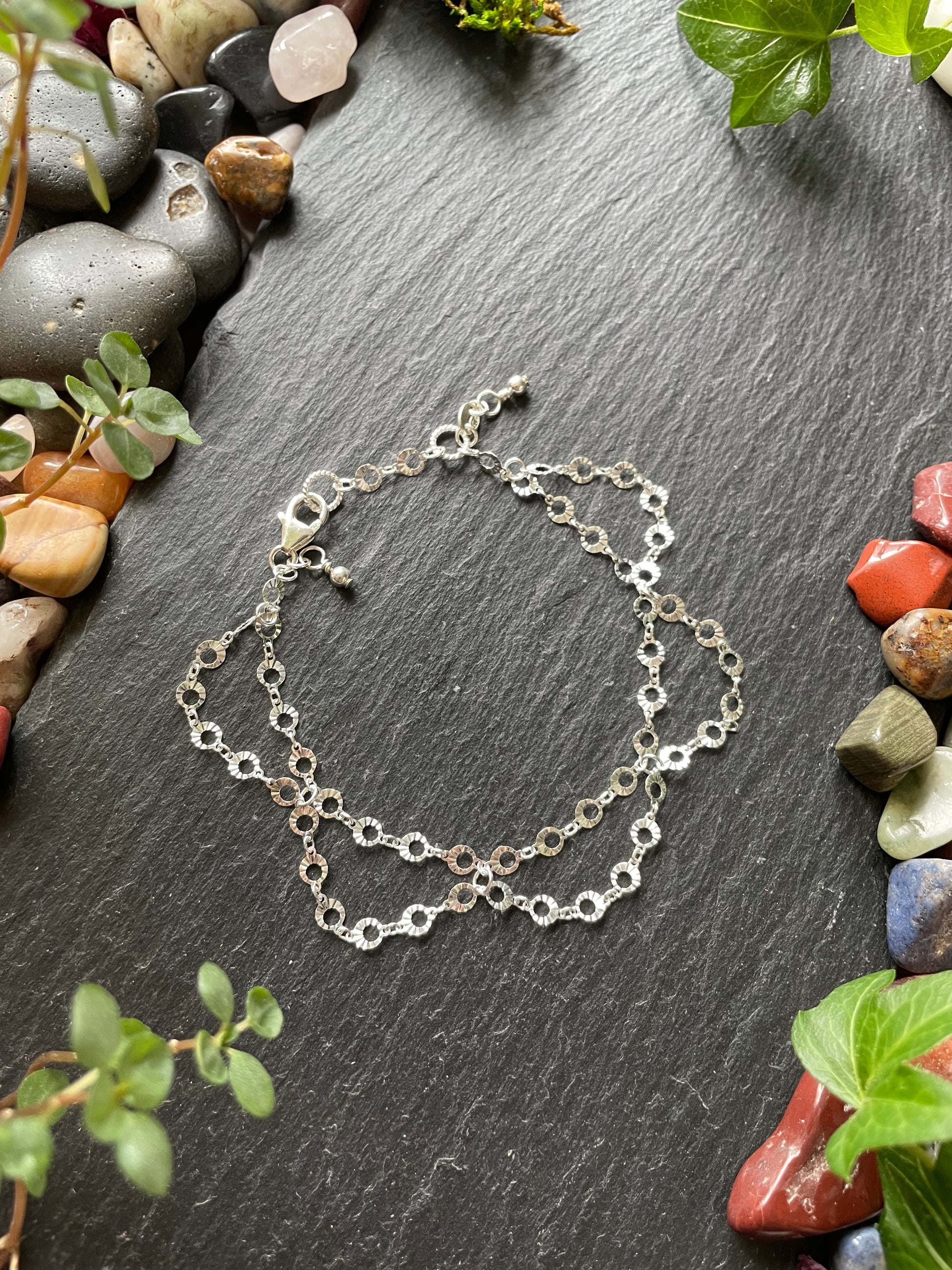 Silver chain bracelet on a dark stone surface with small stones and greenery around