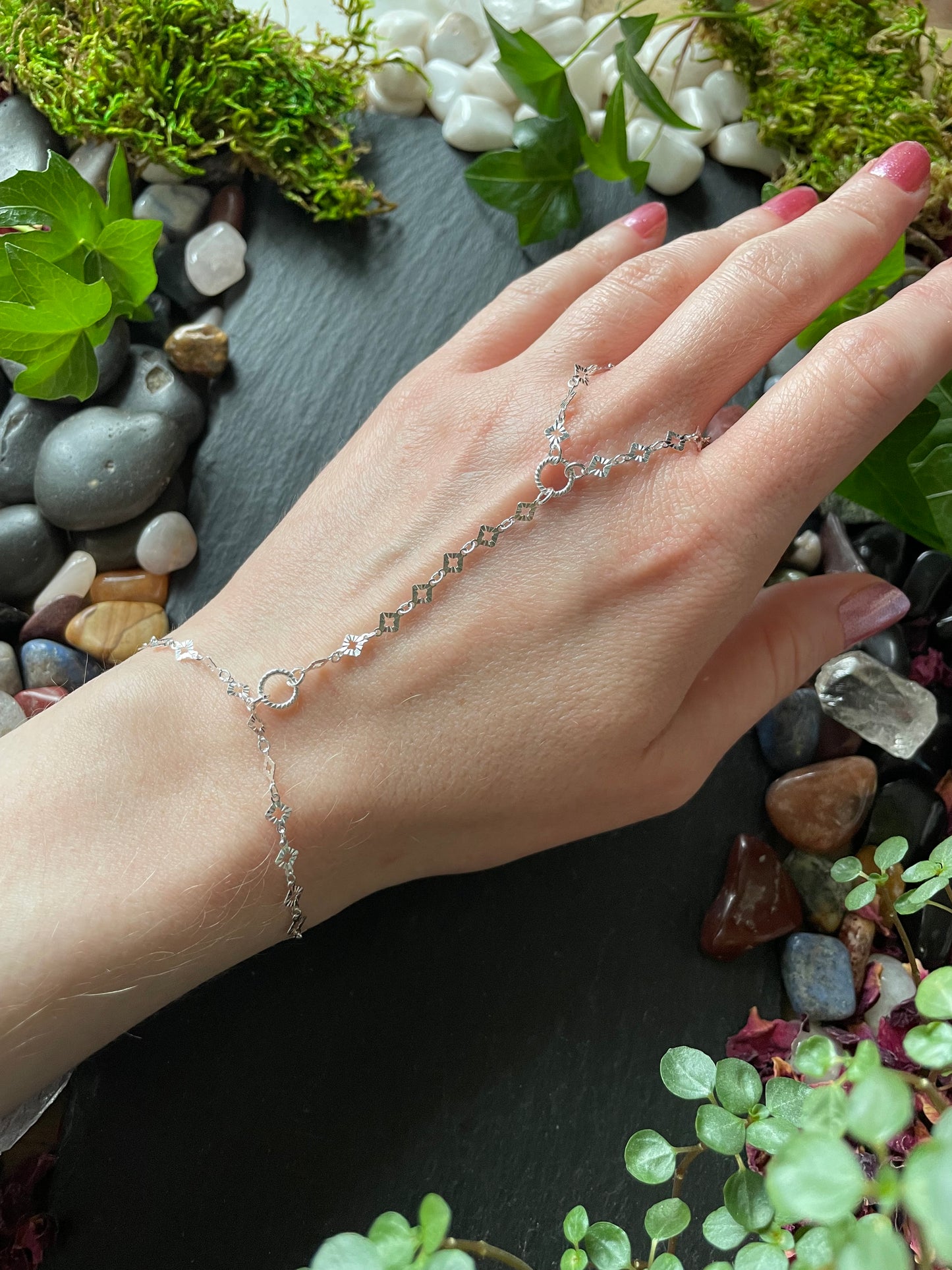 Hand wearing a delicate silver bracelet with a natural stone and plant background