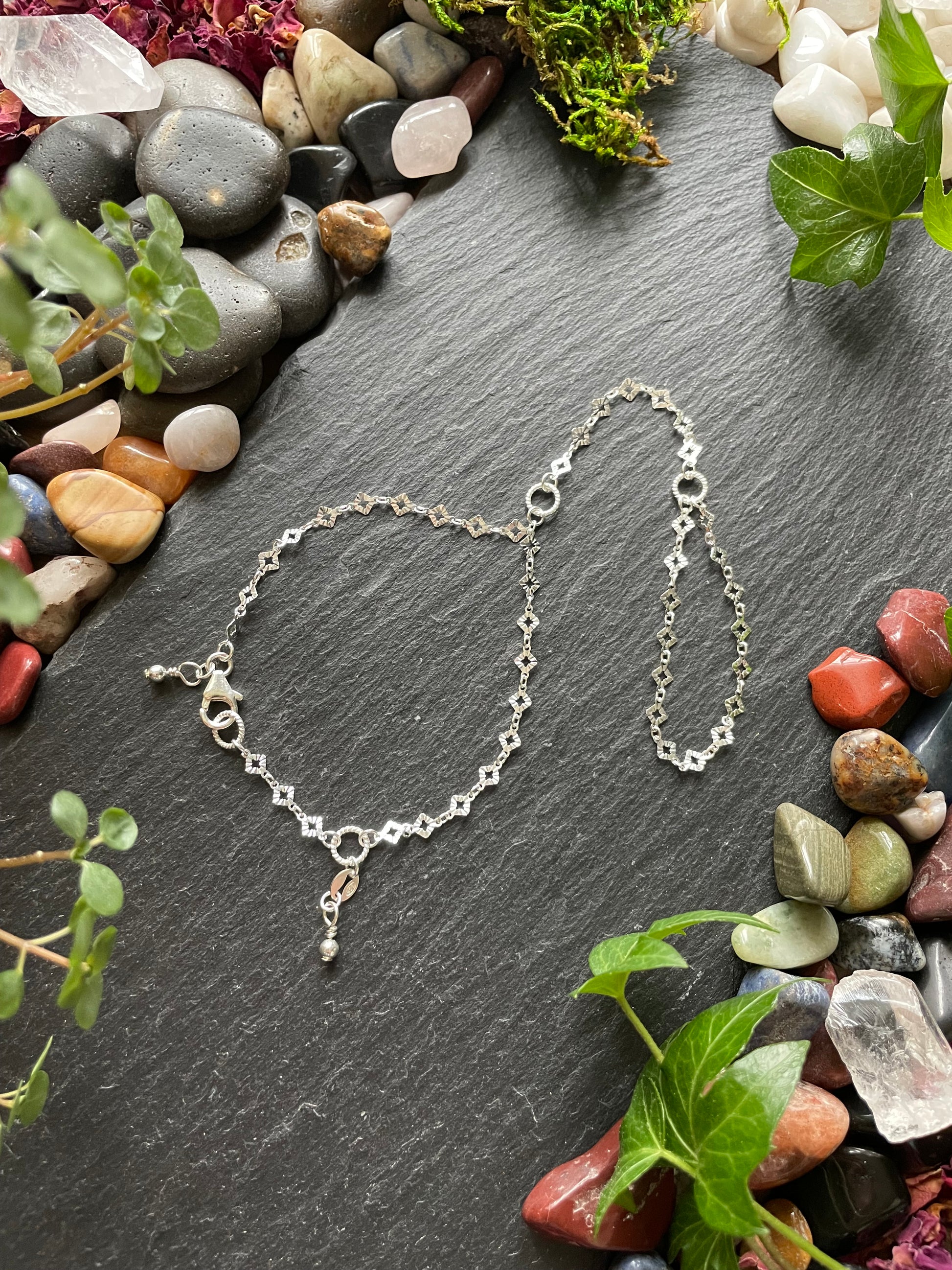 Silver hand chain bracelet on a dark stone surface with pebbles and greenery