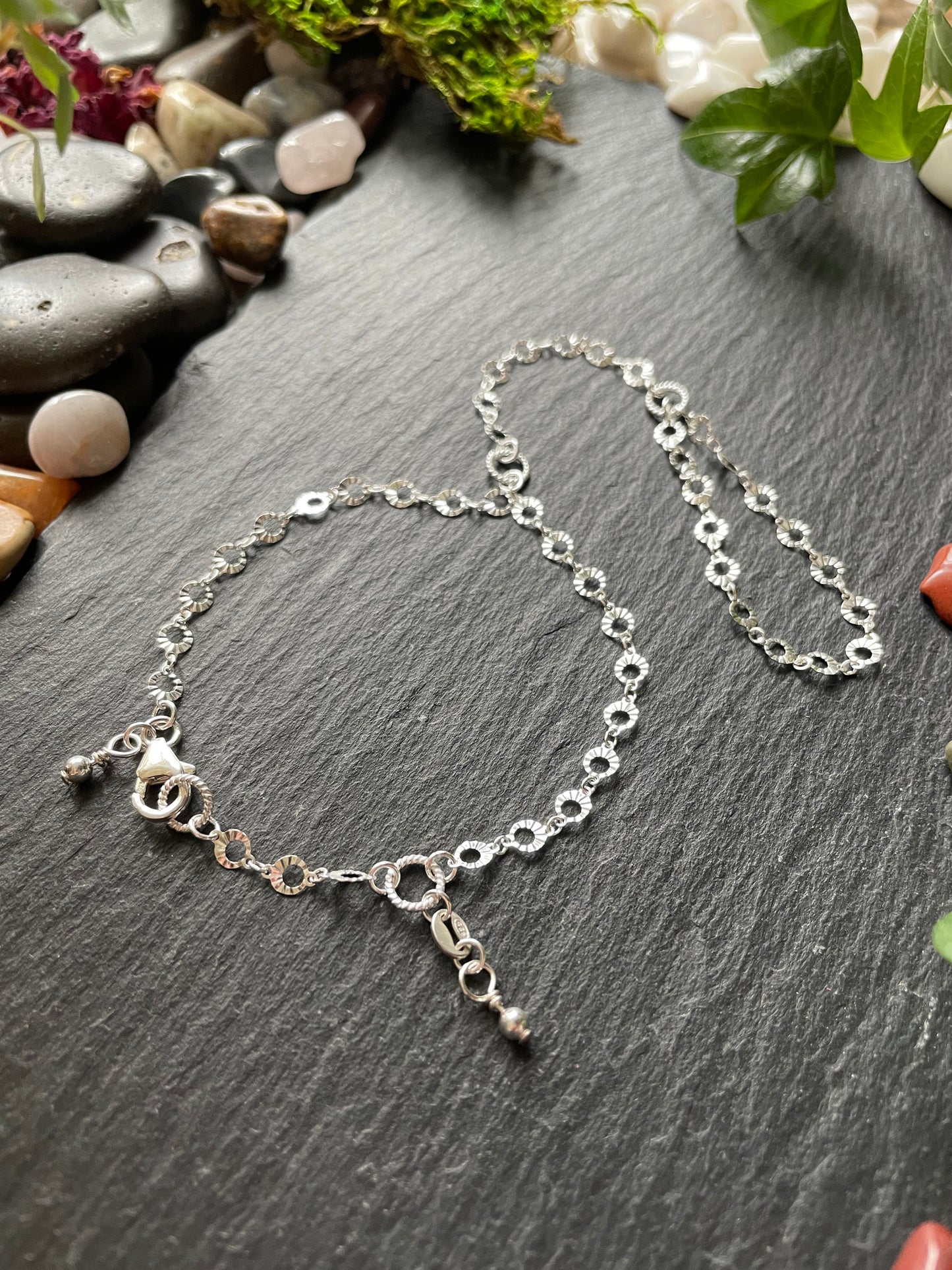 Silver chain bracelet on a dark surface with pebbles and plants in the background