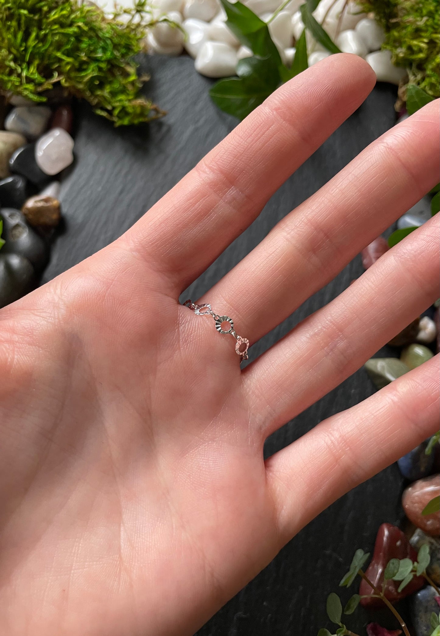 Hand wearing a delicate ring with a natural stone on a textured surface with stones and plants.