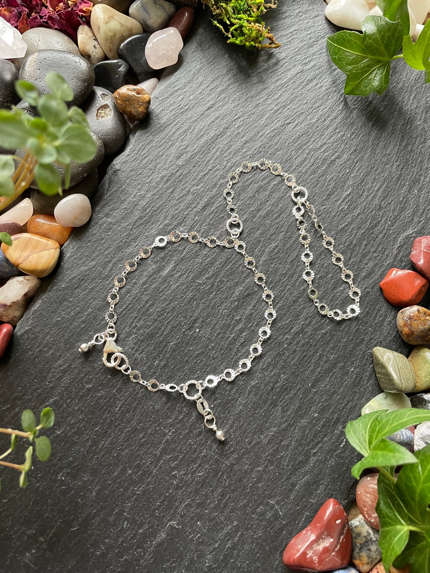 Silver hand chain on a dark stone surface with pebbles and greenery