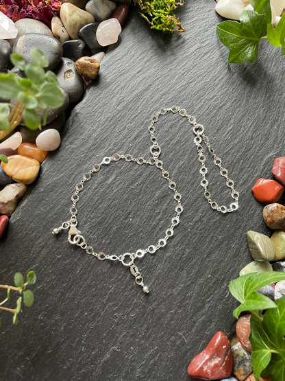 Silver hand chain on a dark stone surface with pebbles and greenery