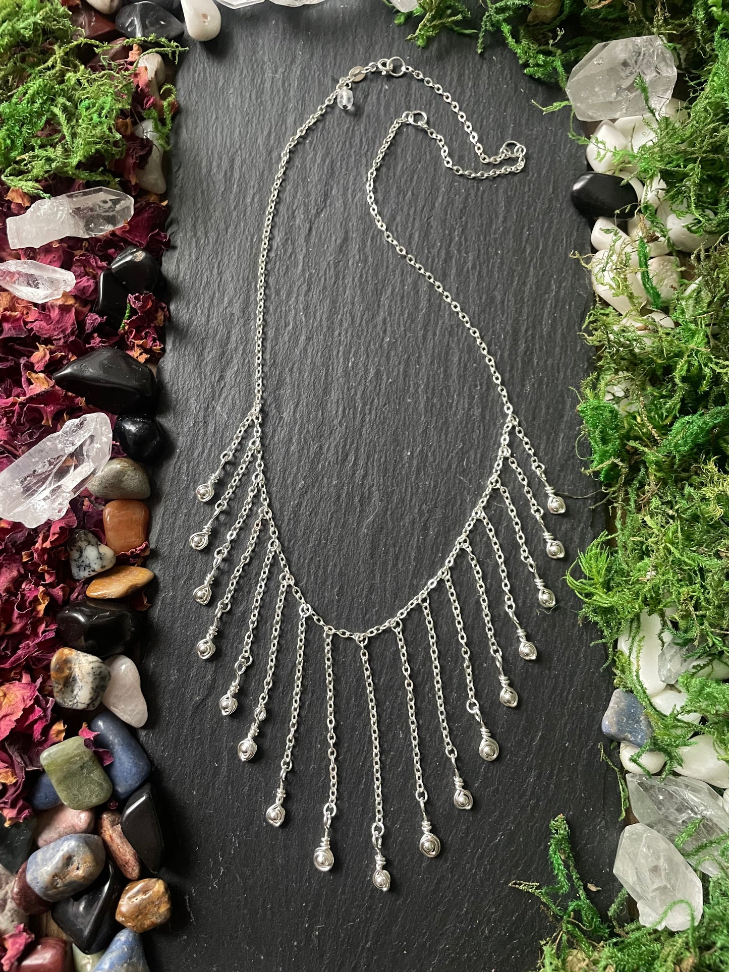 Silver fringe necklace on a dark slate surface with natural elements around