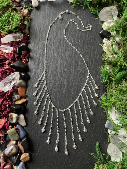 Silver fringe necklace on a dark slate surface with natural elements around