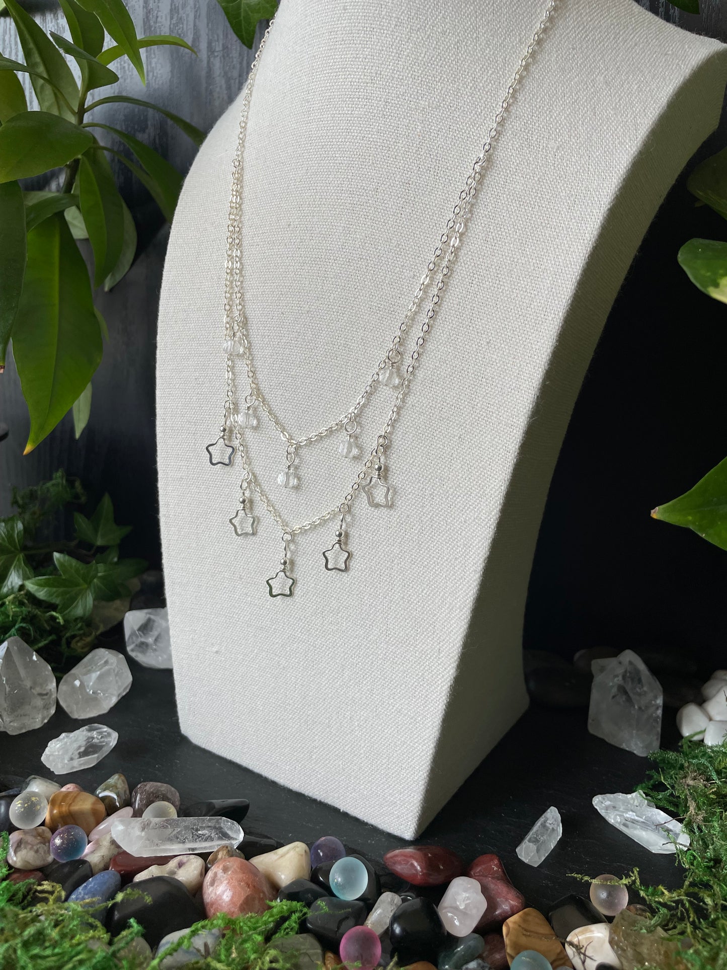 Silver necklace with star charms on a white bust, surrounded by crystals and plants.