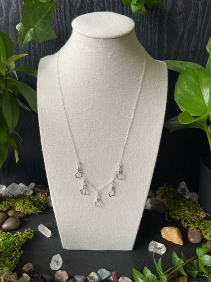White jewelry display with necklace on a black surface surrounded by greenery and small stones.