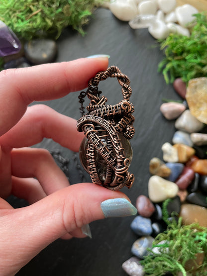 Hand holding a detailed copper wire-wrapped crystal against a natural stone and moss background