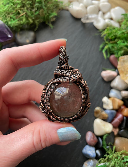 Hand holding a copper wire-wrapped amulet with a crystal against a natural stone and greenery background.