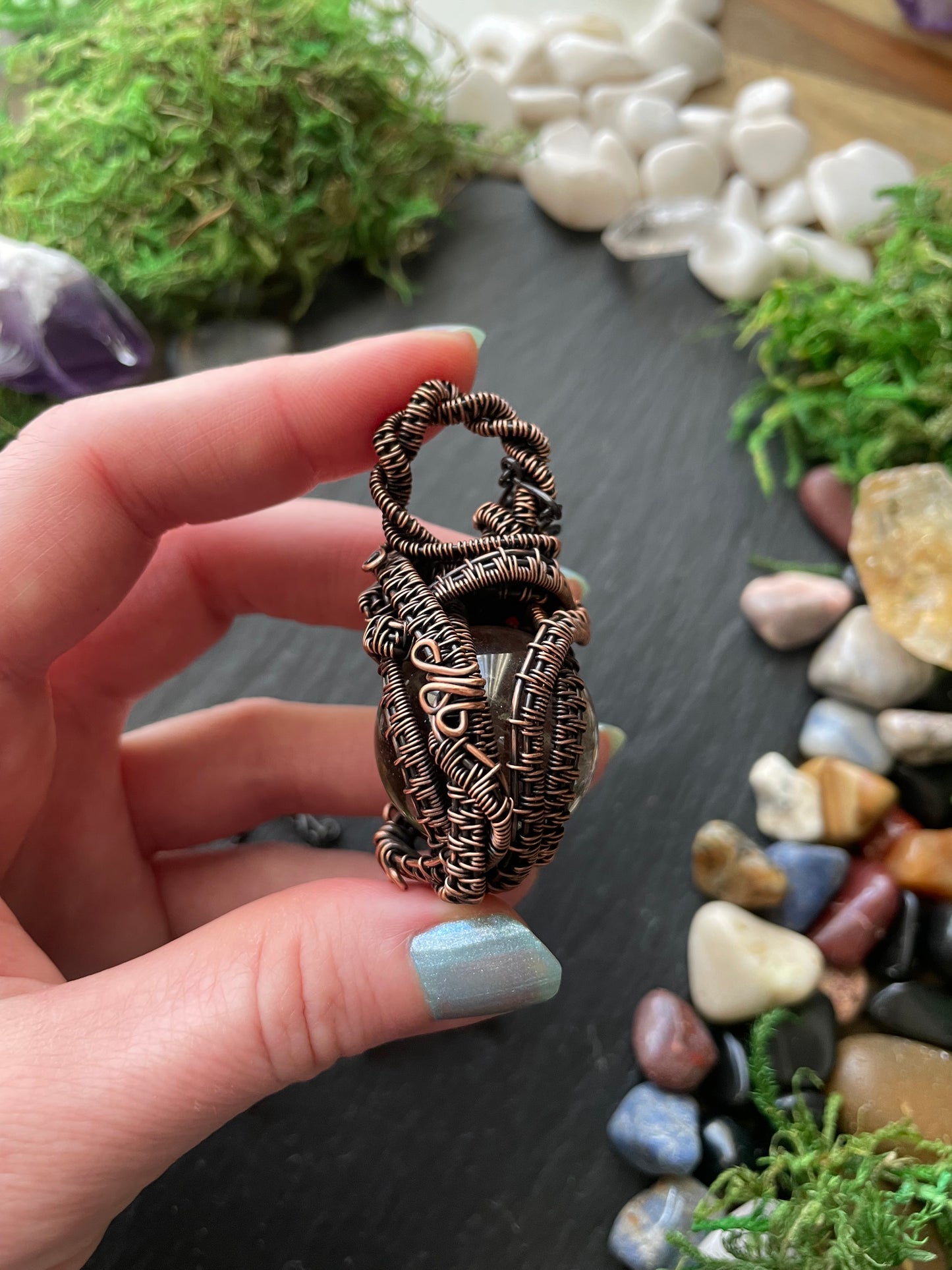 Hand holding a copper wire-wrapped amulet with a natural stone and moss background