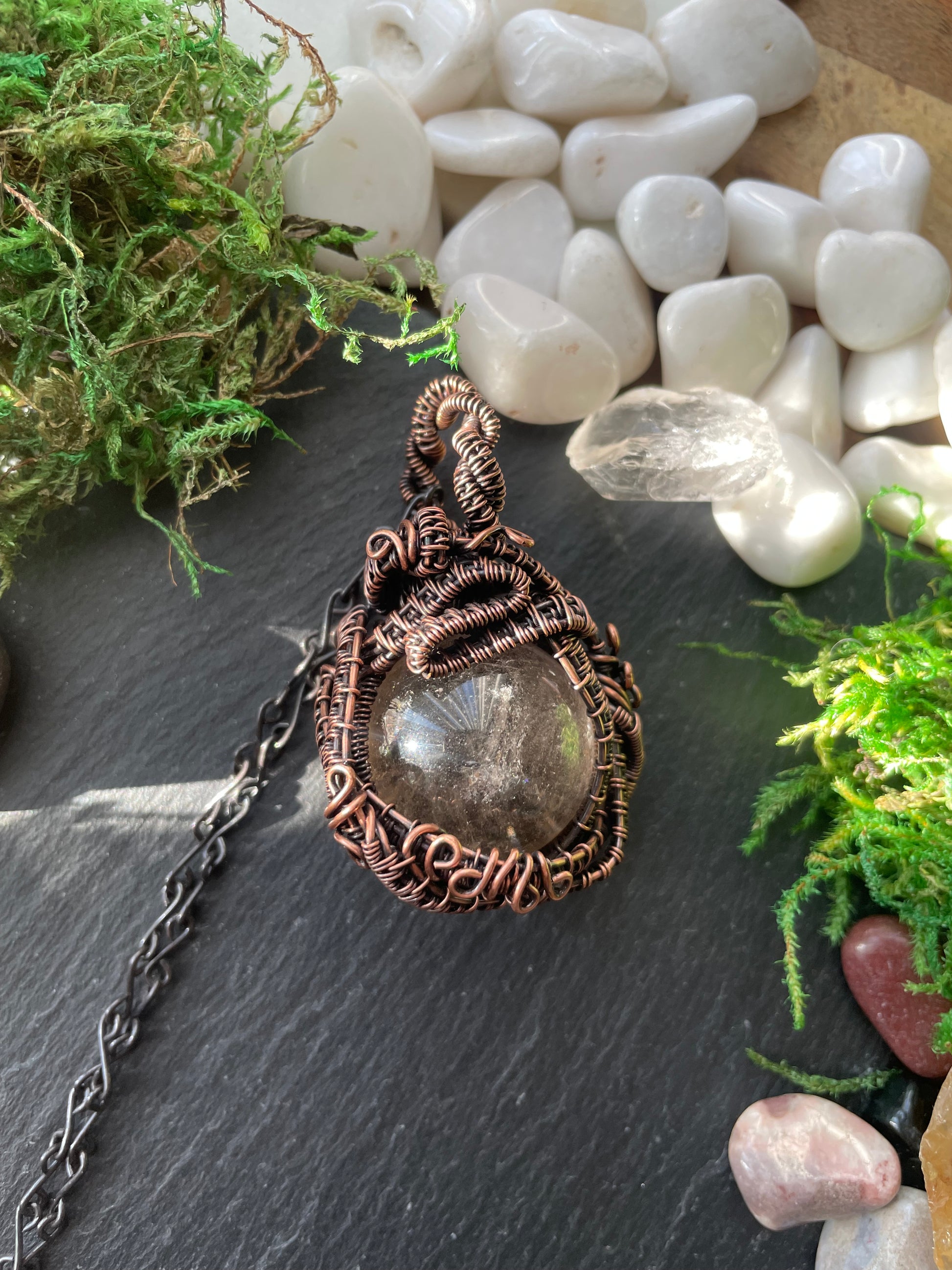 Copper wire-wrapped amulet on a dark surface with pebbles and moss