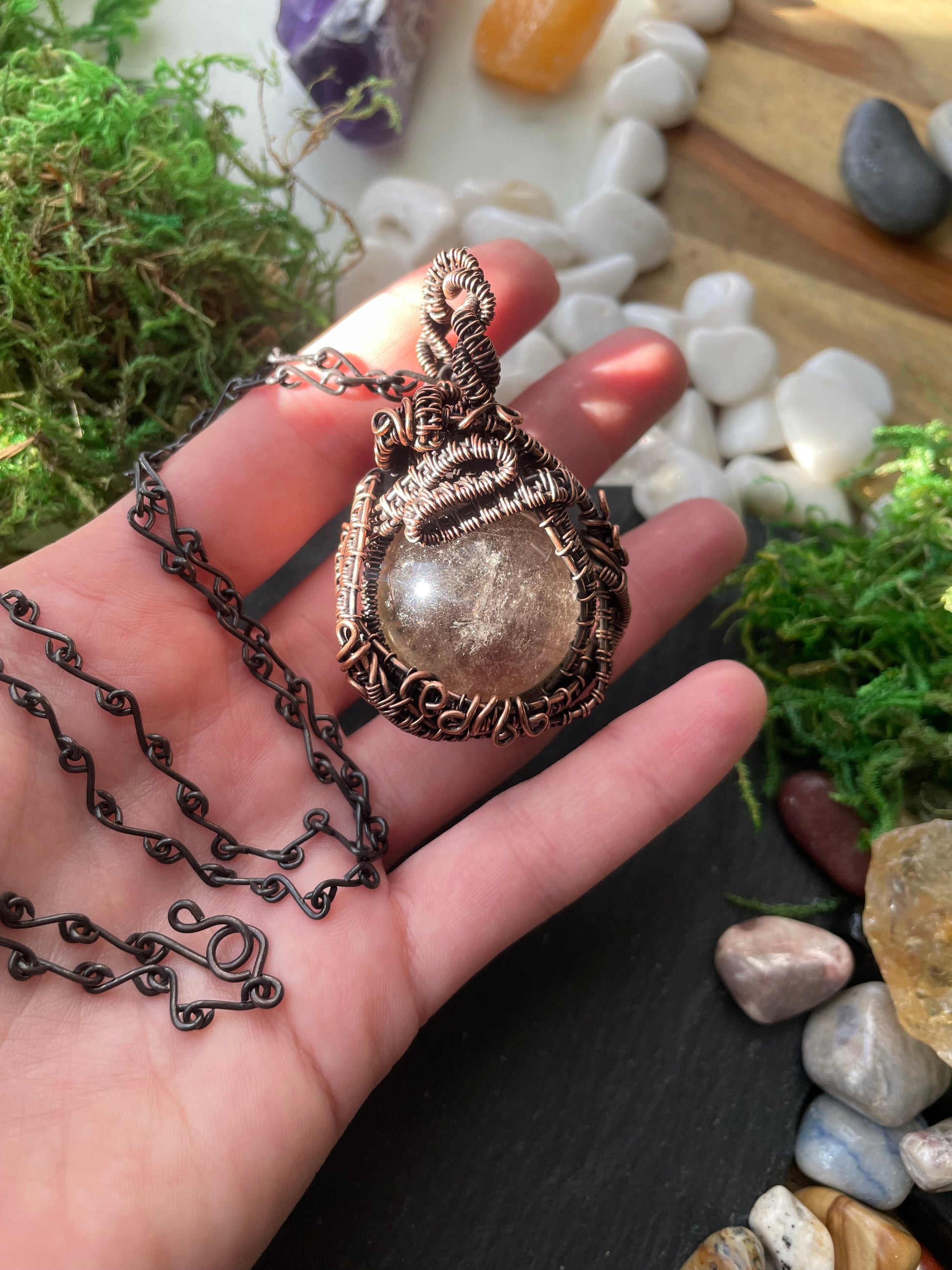 Hand holding a decorative amulet with a chain against a natural stone and moss background