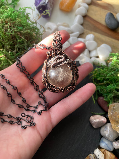 Hand holding a decorative amulet with a chain against a natural stone and moss background