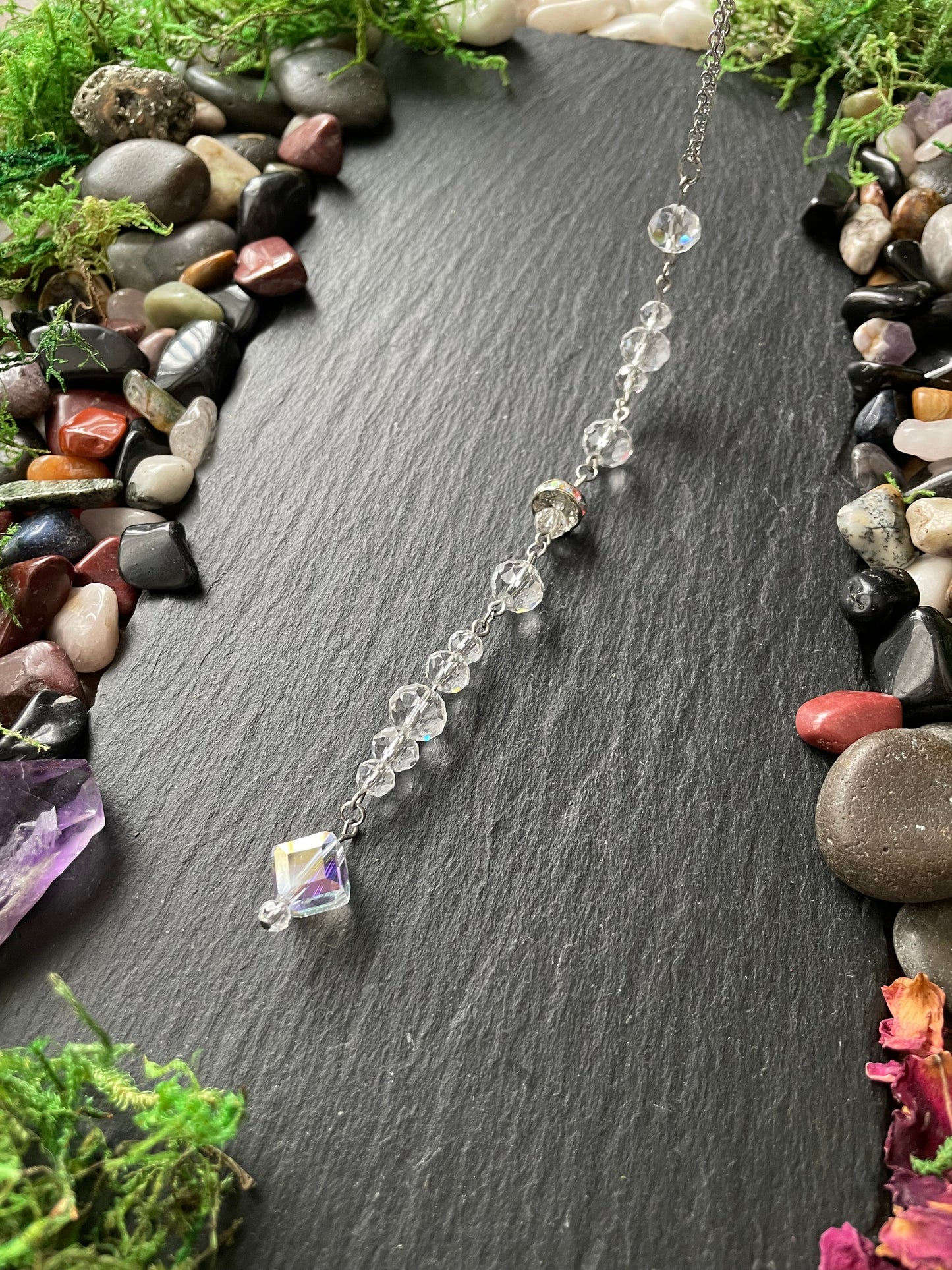 Silver car mirror charm with a clear sparkly glass beads  on a dark stone surface surrounded by gems and plants
