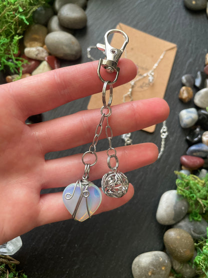Hand holding a silver keychain with heart and crystal cage on a dark surface with stones and moss.