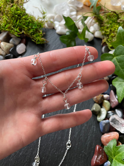 Hand holding a delicate silver necklace with clear beads against a natural stone and greenery background