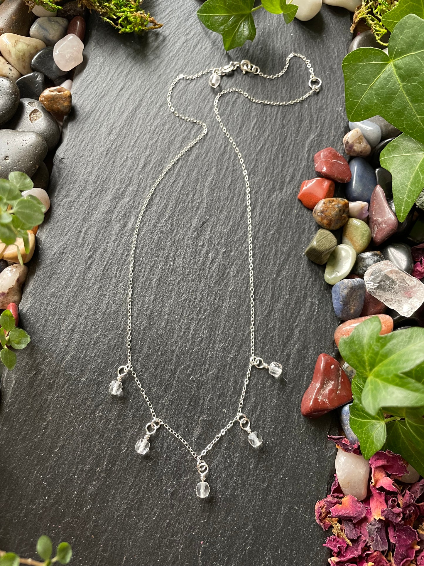 Silver necklace on a dark stone surface with greenery and stones