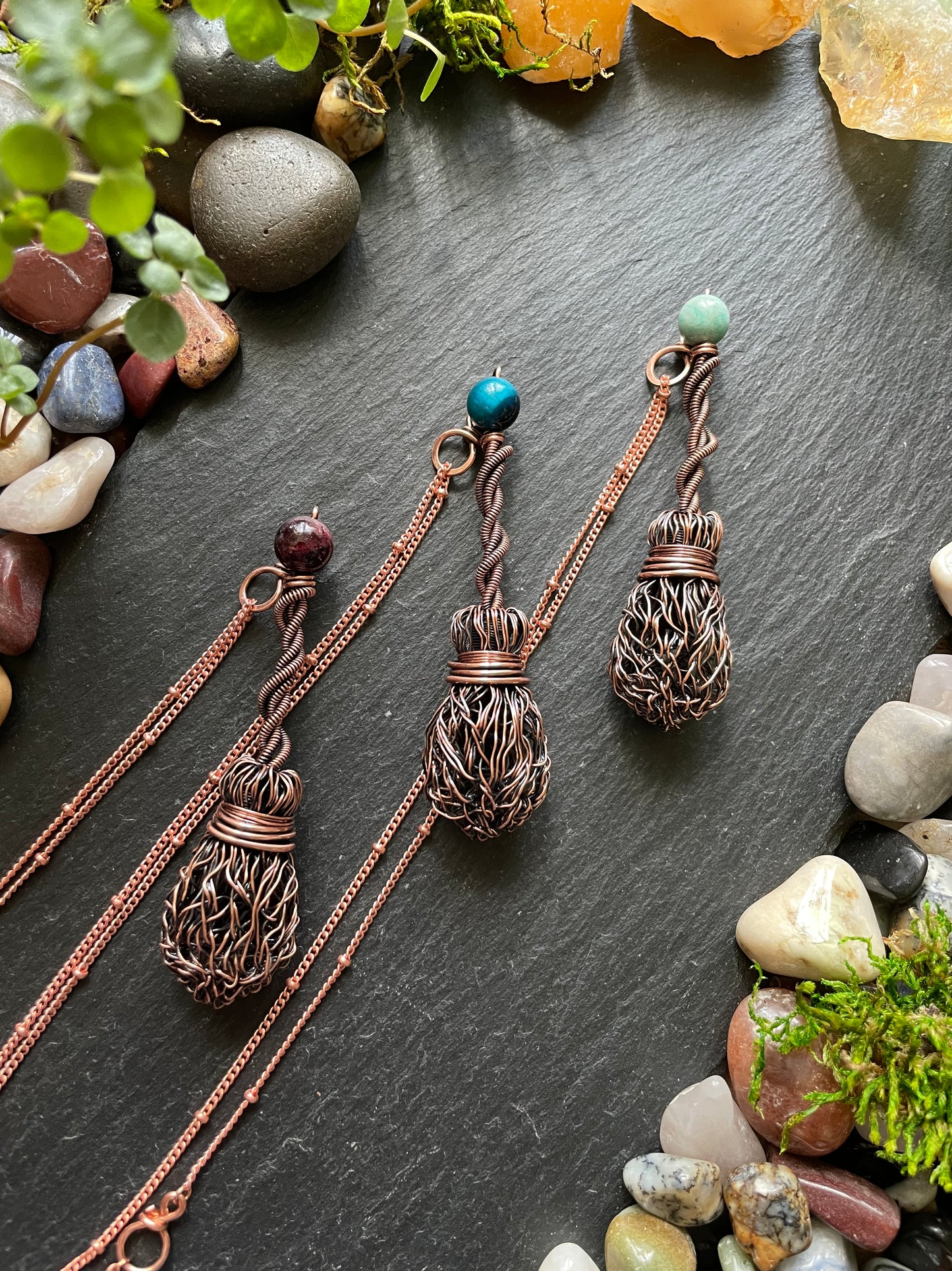 Three copper necklaces with name tags on a dark surface with stones and plants.