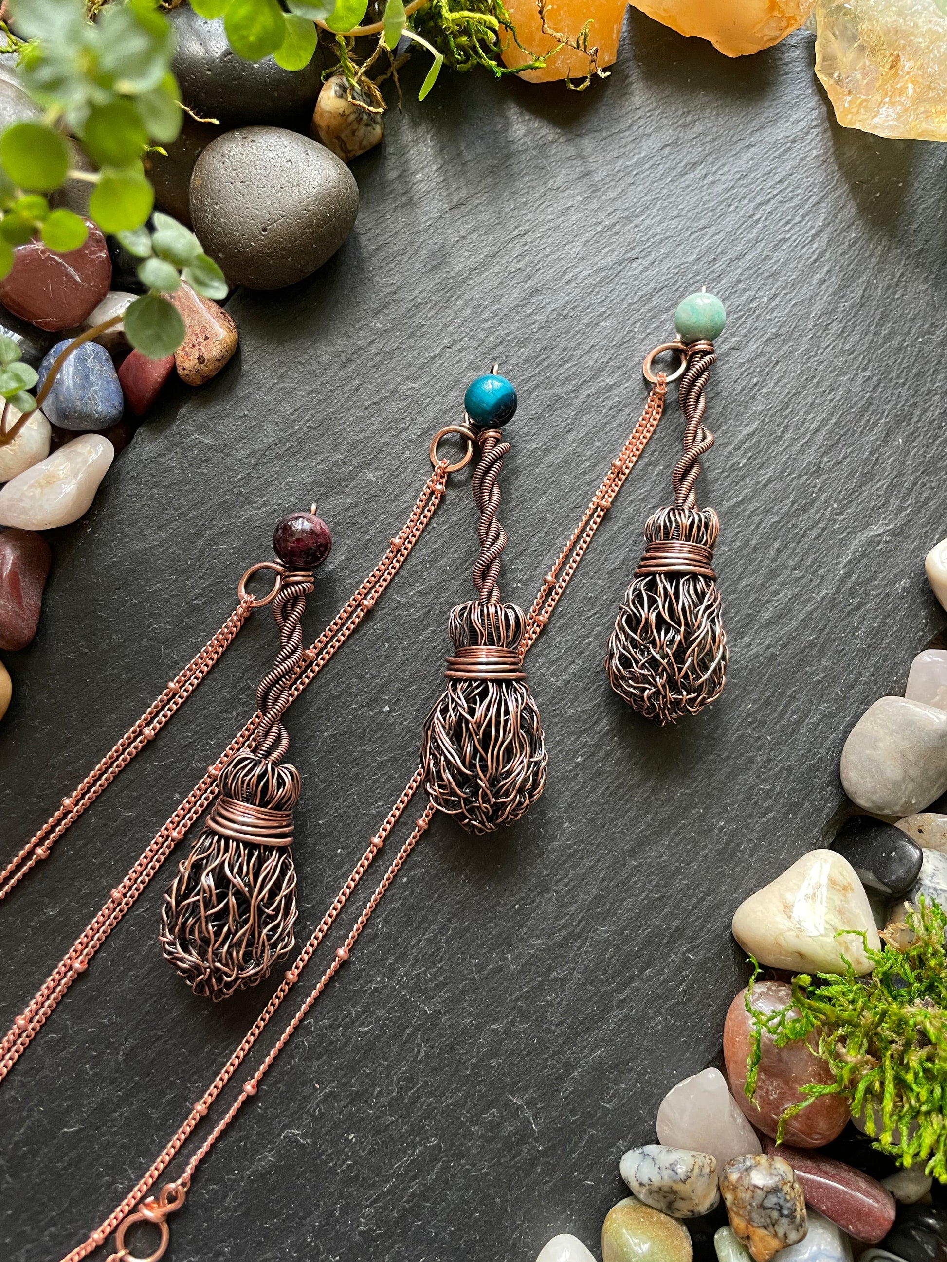 Three copper necklaces with name tags on a dark surface with stones and plants.