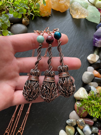 Hand holding three copper brooms with beads against a background of stones and plants